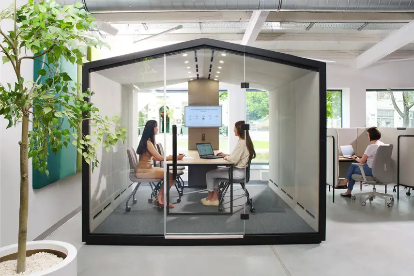 Glass acoustic office pod with two people working at a table, featuring a screen, laptops, and modern seating in an open workspace.